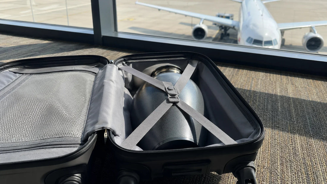 Open suitcase at an airport gate containing a large metallic urn secured with crossed luggage straps, with a passenger airplane visible through the terminal window in the background.