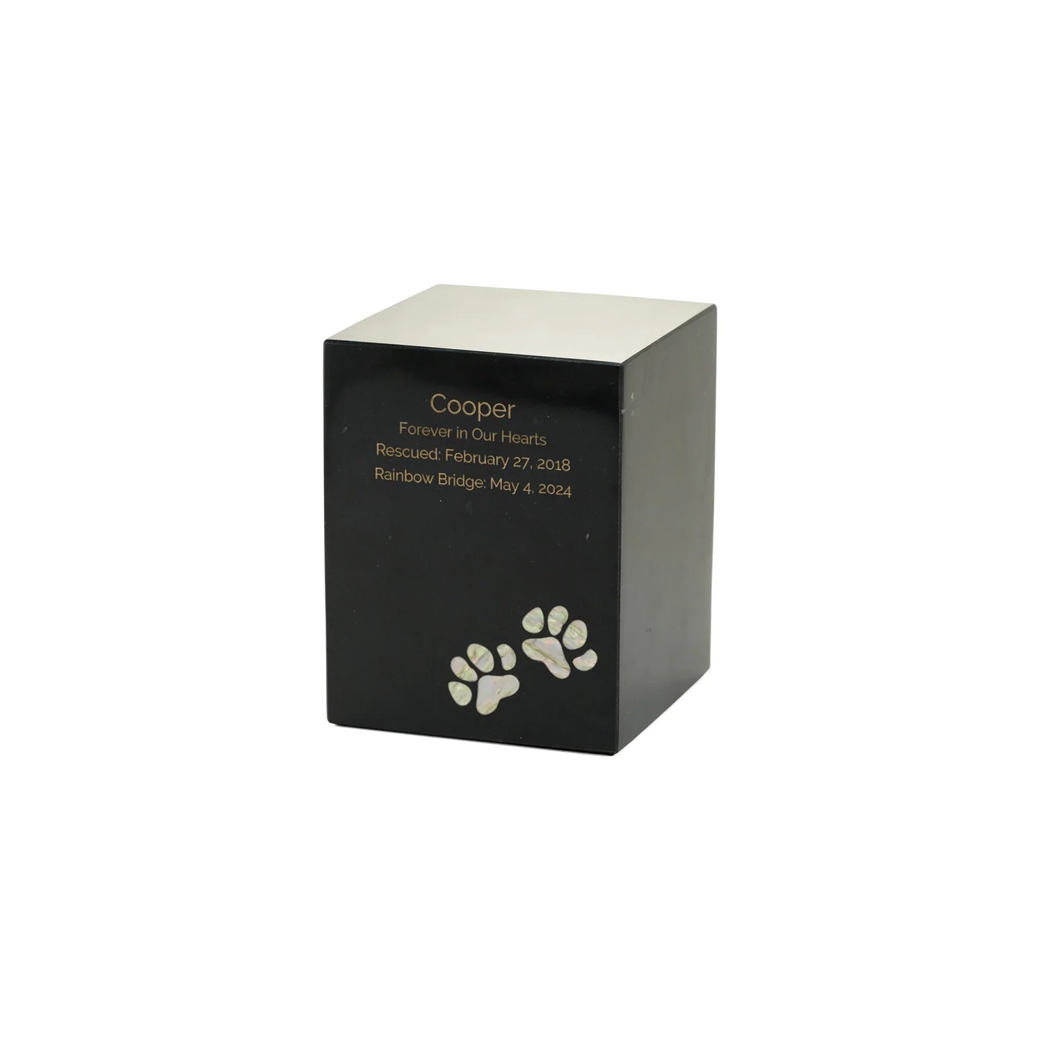 Engraved pet urn featuring a black cube body with a silver top personalized with gold text "Cooper Forever in Our Hearts Rescued: February 27, 2018 Rainbow Bridge: May 4, 2024" and two iridescent mother-of-pearl paw prints