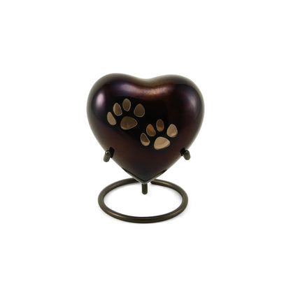 Metal heart-shaped pet urn with a dark iridescent finish and two golden paw prints resting on a minimalist metal stand against a white background